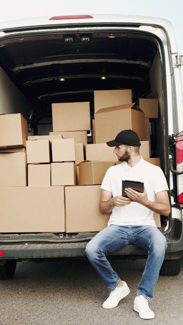 A man wearing a blue beanie, red and navy jacket, black pants, and sneakers stands with arms crossed in front of an open cargo van parked outside a modern building with a white facade and large glass