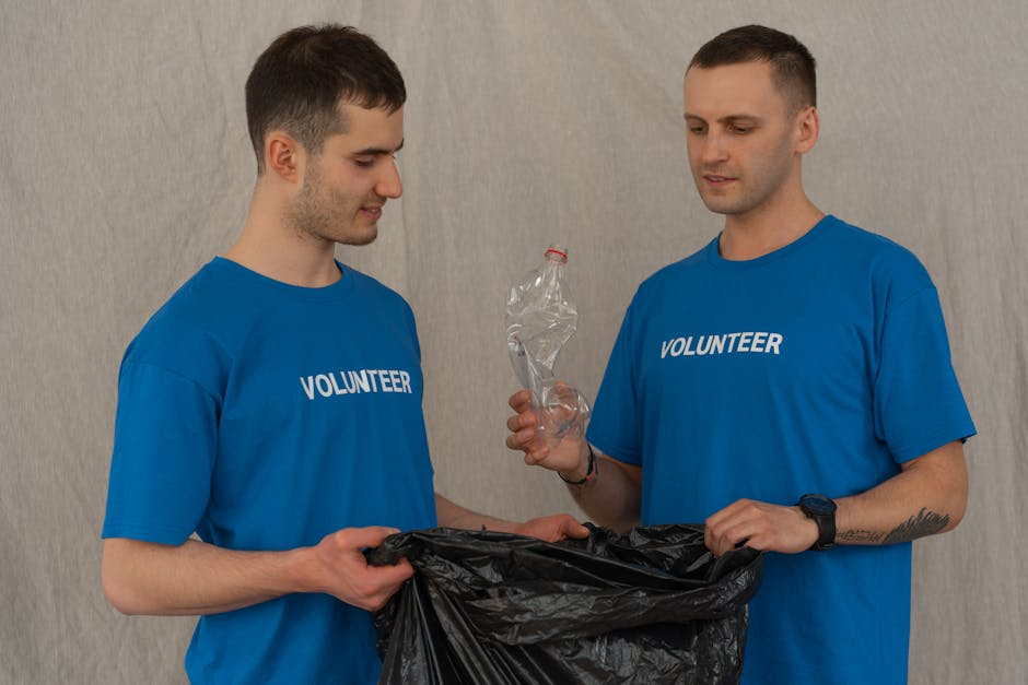 Two male volunteers wearing blue t-shirts with the word 'VOLUNTEER' printed on the front are participating in a home relocation activity indoors against a neutral grey wall. The volunteer on the left