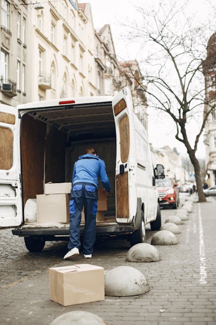 A man wearing a blue jacket and dark trousers is seen loading a cardboard box into the back of a white removal van parked on a city street. The van's rear doors are open, revealing a partially packed