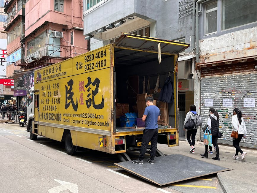 A man loading cardboard boxes onto the back of a white van parked on a residential street during daylight hours. The van's rear door is open, revealing an empty interior prepared for home relocation i
