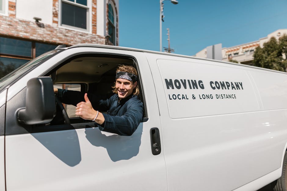 A man with a beard, wearing a black cap, white polo shirt, and wireless earbuds, is seen loading or unloading cardboard moving boxes from the back of a van parked outdoors near a body of water surroun