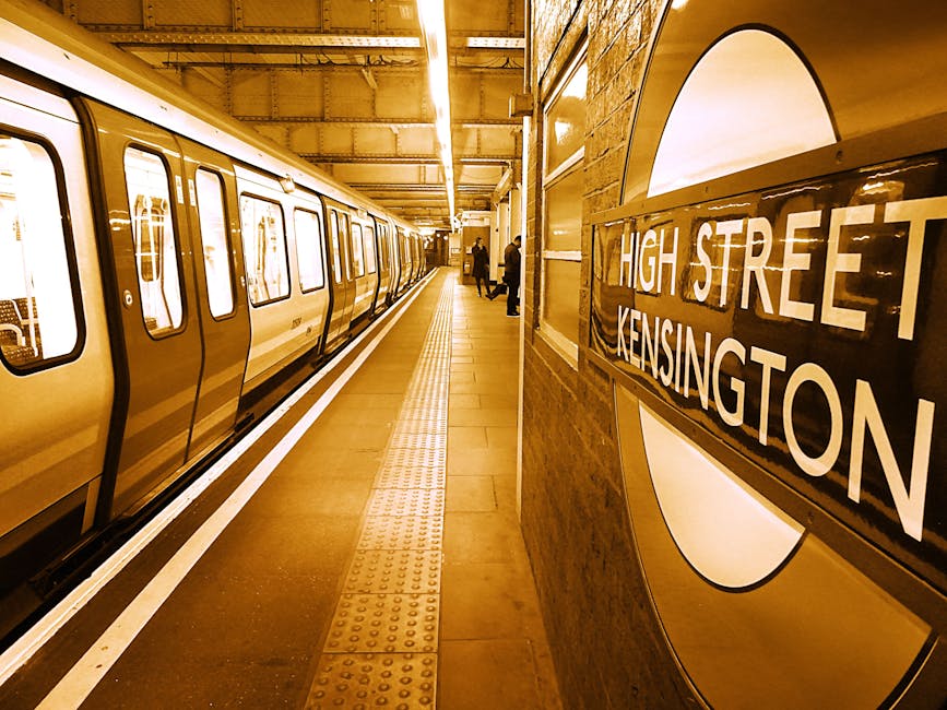 Inside West Kensington underground station, the platform is visible with a yellow train halted alongside. The platform surface is tiled with tactile paving strips near the edge for safety, and a few passengers can be seen waiting or walking in the distance. On the right side, a large round London Underground sign displays the station name 'High Street Kensington' in white letters on a black background, mounted on a curved wall made of brown brick. The station's overhead lighting illuminates the space with a warm, yellowish glow, reflecting off the metallic surfaces of the train and signage. This setting depicts a typical urban transport environment suitable for home relocation or moving logistics, with the visible train carriage and station features highlighting the importance of efficient transport access for furniture transport and packing during a moving process. The clarity of objects, materials, and environment supports accessibility and natural understanding of the scene within a professional removals context, such as those offered by Man with Van West Kensington.