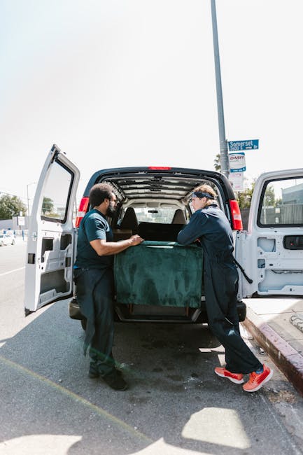 Two movers, dressed in dark uniforms, are loading a large, wrapped piece of furniture into the back of a black van, parked on a street with a curb and a signpost visible in the background. The furniture appears to be protected with green fabric or padding to prevent damage during transport. The open van doors reveal the spacious cargo area, with the interior designed for home relocation tasks, including space for boxes, appliances, and furniture. One mover is holding the furniture steady while the other assists in guiding it into the van. The scene takes place under clear daylight, highlighting the professional process of furniture transport as part of a house removal or moving service. The location suggests a narrow street or access point, relevant to urban removals, aligned with the West Kensington station access tips and associated with Man with Van West Kensington's removal services.